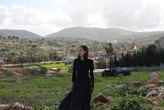 A medium shot of a woman with dark, shoulder-length hair wearing a long-sleeved black top and a long black skirt. She stands in a grassy green field, looking thoughtfully off-camera toward the right. In the background, a sprawling hillside village with tightly packed concrete buildings extends across rolling hills under a bright, overcast sky. A red tractor with a blue water tank trailer is parked in the mid-ground.