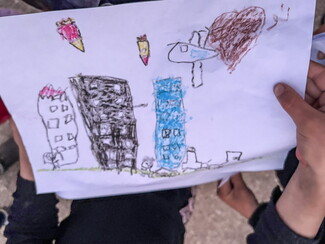 A child's drawing showing a city scene under war, featuring falling rockets, a military plane, a large brown heart, and damaged buildings.