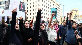 A crowd of people protest in a city street, raising their fists and shouting; one person holds a sign with Arabic writing and a photo of a man.
