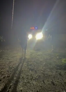 A dark, grainy nighttime photograph showing the blinding headlights and flashing red and blue lights of an emergency vehicle parked on a rough, unpaved surface. Silhouettes of people can be seen walking through the bright glare.