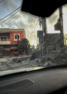 A view from inside a car looking through the windshield at a street covered in rubble, debris, and thick dust or smoke following a strike. A moderately damaged orange multi-story building stands on the left side of the street.