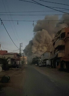 A street-level view in a residential neighborhood where a massive, towering plume of thick grey smoke rises high into the sky from directly behind the buildings on the right side of the road. A string of bare light bulbs hangs across the street under a dim sky.