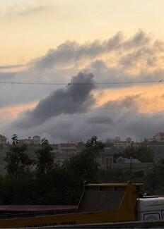 A view of a hilly residential area under a sky filled with massive, dark, billowing smoke clouds tinged with an orange glow from the rising sun. In the foreground, the yellow bed of a Mercedes-Benz dump truck is visible behind some trees.