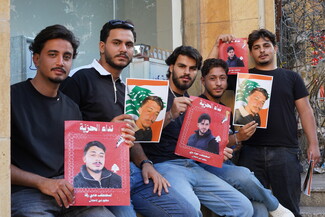 Five young men sit outside a building next to a barbed-wire fence, holding red and Lebanese-flagged protest posters featuring portraits of missing loved ones. The posters feature portraits of the missing men, Lebanese flags, and Arabic text reading “Call for Freedom.”