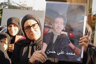 A woman in a black hijab and glasses holds up a large, laminated portrait of a missing man named Mohammad Ali Hassan during an outdoor demonstration. Other women stand closely behind her in solidarity.
