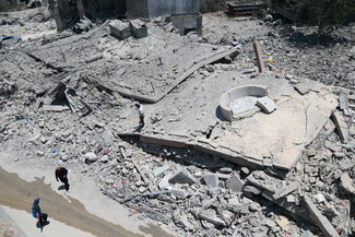 A high-angle view looking down at a large concrete multi-story building that has completely pancaked flat onto the ground in Bir el-Salasel, South Lebanon. Three men stand or walk on the cleared asphalt street adjacent to the massive slabs of flattened concrete.