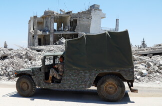 A side profile view of a camouflage military vehicle with a dark green soft-top canvas being driven by a soldier in uniform. In the background across the street stands a heavily shelled, ruined multi-story concrete building with its interior rooms completely exposed.