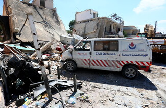 A white Islamic Health Association Civil Defense van with red striped markings and Arabic text reading "Srifa town" parked next to a severely damaged building that has pancaked diagonally. Rubble, household debris, and twisted metal cover the ground, with a yellow excavator visible working in the background.