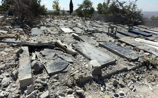 A severely damaged cemetery in Srifa, South Lebanon. The black and gray marble tombstones are cracked, broken, and heavily blanketed in concrete dust, loose rubble, shattered blocks, and uprooted tree branches. A stray mattress lies discarded among the graves.