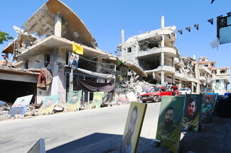 A street-level view of a road in Srifa, South Lebanon, lined with heavily shelled and collapsed buildings. Large commemorative portrait posters of young men line the lower right side of the road. Yellow and green flags hang from wires above the street as a red pickup truck and other vehicles drive through.