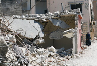 A heavily damaged residential building in Tebnine, South Lebanon, with its facade peppered by shrapnel marks. In the foreground, concrete structures have collapsed entirely into a chaotic mound of rubble and tangled iron reinforcement bars. A woman wearing a black abaya and blue hijab stands on the debris-strewn dirt road on the right, looking toward the camera.