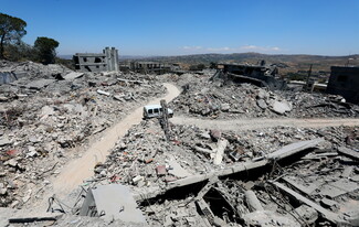 A wide, high-angle view capturing an extensive landscape of devastation in Tebnine, South Lebanon. Entire neighborhoods are reduced to sprawling fields of gray concrete rubble and pulverized buildings. A narrow dirt track snakes through the center of the debris field, where a solitary white minivan is driving under a clear blue sky.