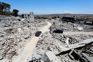 A high-angle panoramic view of massive structural destruction in Tebnine, South Lebanon. Mounds of shattered concrete and exposed rebar line a curved dirt road that has been cleared through the ruins. A dark gray hatchback and a black sedan are navigating the road amidst the completely flattened landscape.