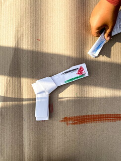 A top-down view of a toy gun folded out of white paper, featuring a hand-drawn Palestinian flag along its barrel, resting on a brown cardboard sheet