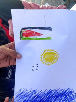 A child's marker drawing on white paper featuring a Palestinian flag, a smiling yellow sun, a small flock of birds, and scribbled blue water at the bottom.
