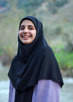 A portrait of a smiling young woman wearing a black hijab and a light purple knitted sweater. She is standing outdoors with a softly blurred background showing greenery and a river.