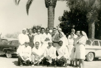 A vintage black-and-white group portrait of about fifteen medical professionals, including Frantz Fanon, among doctors and nurses wearing white hospital uniforms and caps, posing together outdoors. Two classic mid-century cars and large palm trees are visible behind them.