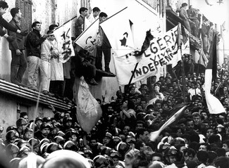 A historic black-and-white photograph of a massive, densely packed street demonstration on the anniversary of the Algerian War for Independence. A line of riot police wearing helmets stands before a crowd of civilians on the street and balconies. Protesters hold up flags and a large white banner reading (French): "ALGERIA WILL LIVE INDEPENDENTLY"