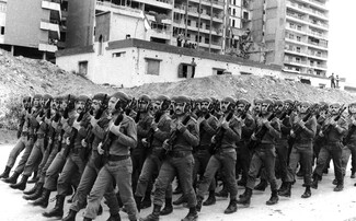 A black-and-white photograph of a large formation of Lebanese Army soldiers marching in tight unison during a drill. The soldiers are dressed in matching uniforms and combat boots, wearing distinctive helmets with integrated ear and chin protection, holding rifles diagonally across their chests. In the background, a war-torn urban landscape features large mounds of dirt and rubble in front of a heavily damaged, multi-story apartment complex and a smaller white building marked with bullet holes.