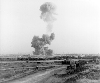 A black-and-white historical photograph capturing a massive, mushroom-shaped smoke cloud billowing high into the sky immediately following the bombing of the U.S. Marine barracks in Beirut on October 23, 1983. In the distant background, the plume rises from the site of the attack near airport buildings. The foreground features an open, flat landscape with a dirt road, a military-style tent, and sandbag fortifications.