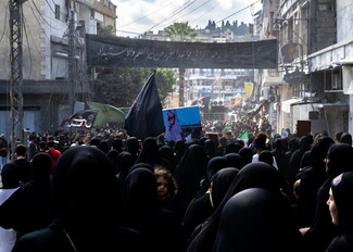 High-angle view from behind a dense crowd of mourners dressed in black walking through an urban street during an Ashoura procession in Nabatieh. Black, green, and blue flags rise above the crowd, beneath a large black banner with white Arabic calligraphy spanning between multi-story buildings.