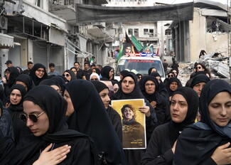 A close-up of women wearing black abayas marching in an Ashoura procession in Nabatieh, resting their right hands on their chests in mourning. In the middle of the group, a woman holds a framed portrait poster of a young man, with an emergency ambulance and war debris visible in the background.