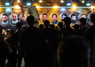 Low-light scene showing the back silhouettes of men gathered before a brightly illuminated memorial wall displaying a row of portraits of young men under overhead lights during an evening Ashoura event in Nabatieh.