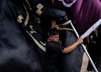 A young man wearing a black shirt strains as he hoists a large metal pole carrying heavy black and maroon religious banners trimmed with gold Arabic calligraphy during Ashoura commemorations.