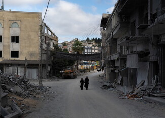 Wide shot of a heavily destroyed street in Nabatieh, flanked by collapsed concrete structures and piles of debris. Two women clad in black abayas walk side-by-side down the empty dusty road beneath a black banner hanging overhead.