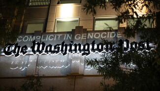 A projection of text reading 'COMPLICITY IN GENOCIDE' is displayed on the facade of The Washington Post building at night, with Arabic text below
