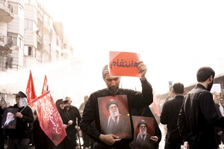 A man dressed in black walks through a crowd holding a framed portrait of Ayatollah Ali Khamenei against his chest while raising a red sign with the Arabic text "#انتقام" (#Revenge) in the air. Red flags and other mourners carrying portraits are visible in the background.