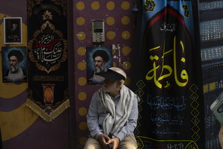 An elderly man wearing a black sun visor, a keffiyeh, and a plaid shirt sits against a wall decorated with black and blue religious banners featuring Arabic calligraphy and posters of Ayatollah Ali Khamenei.