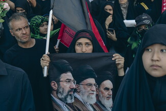 A close-up of a crowd where a woman in a black chador holds a banner depicting three Iranian Supreme Leaders — Mojtaba Khamenei, Ali Khamenei, and Ruhollah Khomeini — surrounded by other attendees.