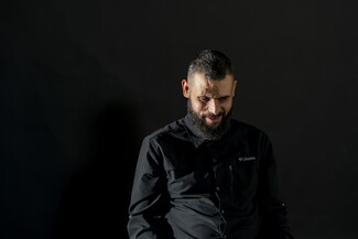 يجلس رجل أمام خلفية سوداء؛ له لحية ويرتدي ملابس سوداء، وينظر إلى الأسفل ضاحكاً بخجل.