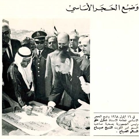 A page from a brochure in Arabic reading "Laying the foundation stone" with an image of President Helou and a smiling Sheikh Al-Sabah as they place their fists on the foundation stone. Dignitaries, at least one in military attire, many in sunglasses, crowd around the leaders, expectantly. 