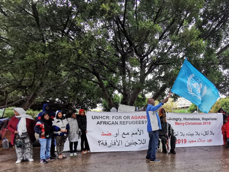 A small group of adults and children stand around a banner that reads, "UNHCR: For or Against African Refugees?" on one side and "Hungry, Homeless, Hopeless: Merry Christmas 2019" on the other. In the center of the image, one man holds the UN Refugee Agency flag. 
