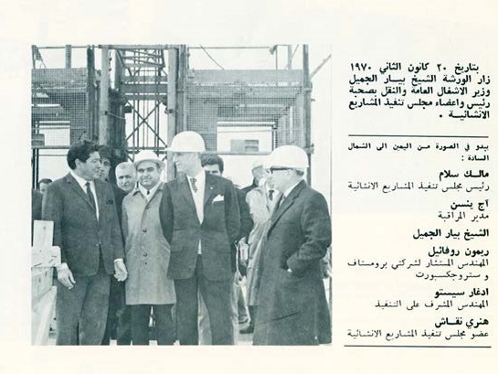 Page of a brochure in Arabic announcing the visit of the Minister of Public Works and Transport Pierre Gemayyel, pictured wearing a hard hat and a three-piece suit with a pocket handkerchief, as he speaks with the head engineer. A group of engineers and officials stand by.