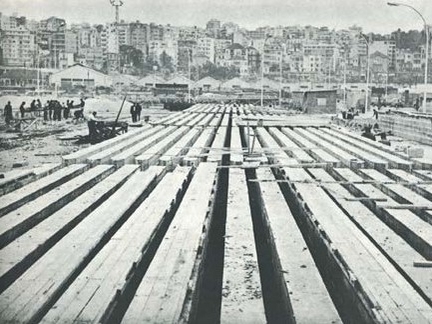 Black and white image of rows of vertically-arranged reinforced concrete piles with the Beirut skyline in the background and workers on the left side 