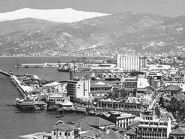 A black and white image of the port viewed from afar. Snowy Mount Sannine appears in the background, the silos are in the center of the image, and the rest of the port apparatus appears in the foreground.