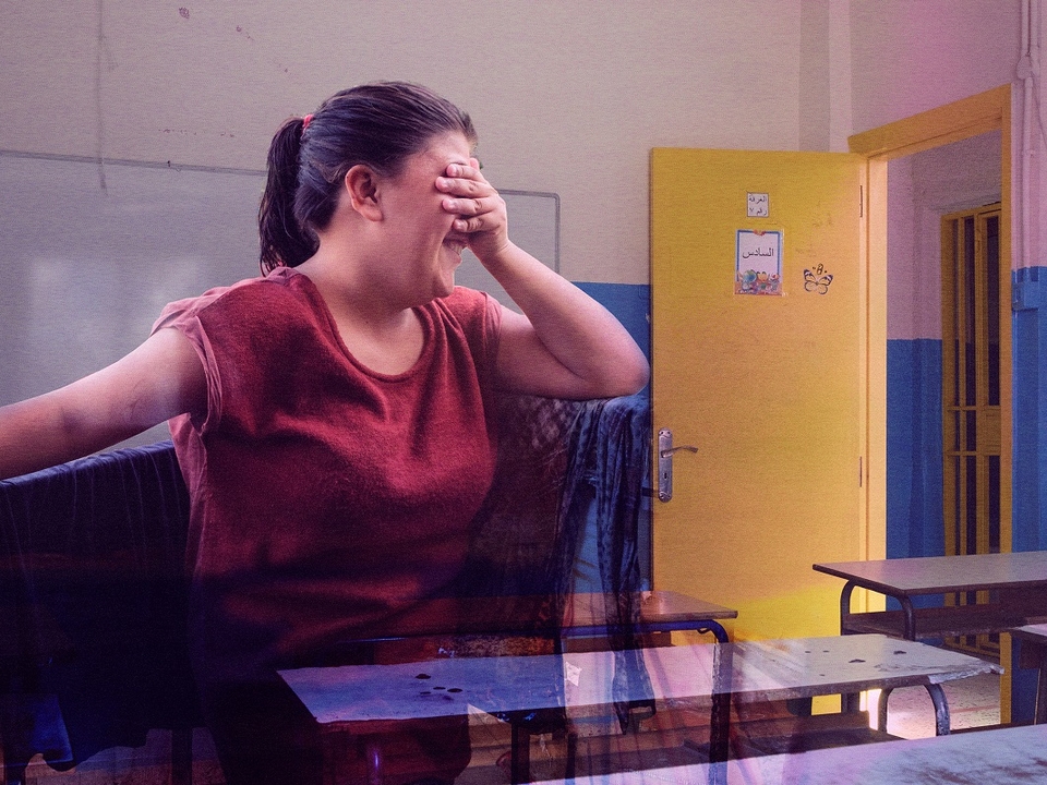 A composite of two images: a girl covering her eyes and looking away with a shy smile on her face in the foreground, with a classroom in the background.