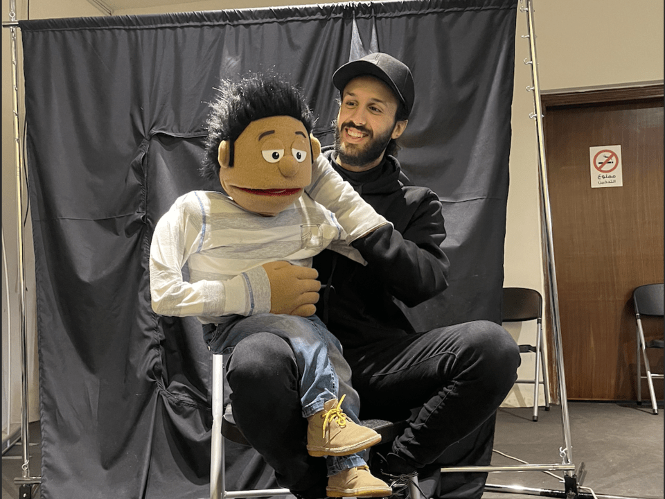 A smiling young man dressed in black is sitting on a chair and holding up a puppet.