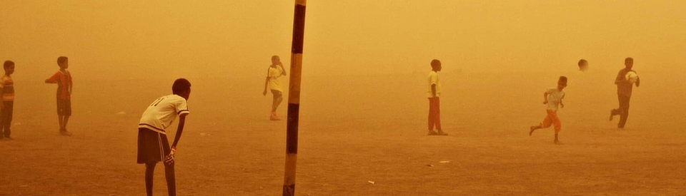 Kids playing football on the sand in the middle of a sandstorm that colors the entire photo golden-yellow.