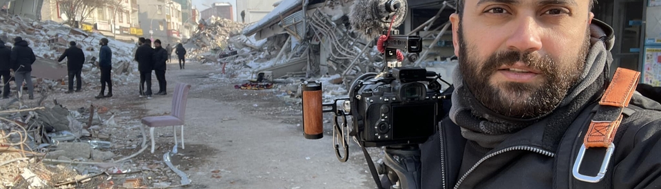 A self-portrait of journalist Issam Abdallah in the foreground, carrying his DSLR camera and microphone, to the background of destroyed buildings. Clear winter day.