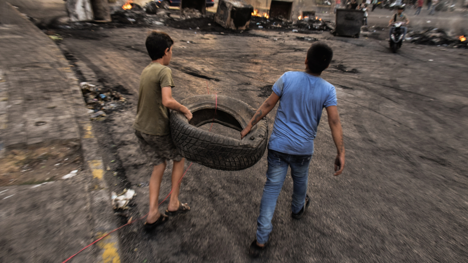 طفلان يحملان دولابا عند حاجز في الطريق مقطوع بمستوعبات النفايات المشتعلة. بيروت، لبنان. ١٨ تشرين الأول ٢٠١٩. (موران مطر، فورة)