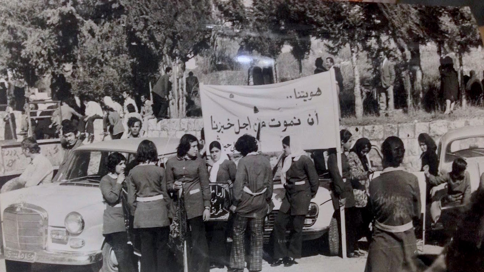 Young women in factory uniforms hold a banner that reads, “our identity is to die for our bread.” Date unknown. Source: Al-Anwar newspaper.     