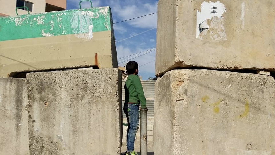 طفل يقف ضمن فتحة ضيقة بين مكعبات اسمنتية. يحدد الجيش اللبناني مرور السيارات إلى عدد من المخيمات الفلسطينية عبر حواجزه ويغلق المداخل الأخرى بجدر اسمنتية (سواتر) فيها ممرات للمشاة فقط. مخيم برج الشمالي، صور. ٢١ كانون الثاني ٢٠١٧. (ناديا أحمد، مصدر عام)