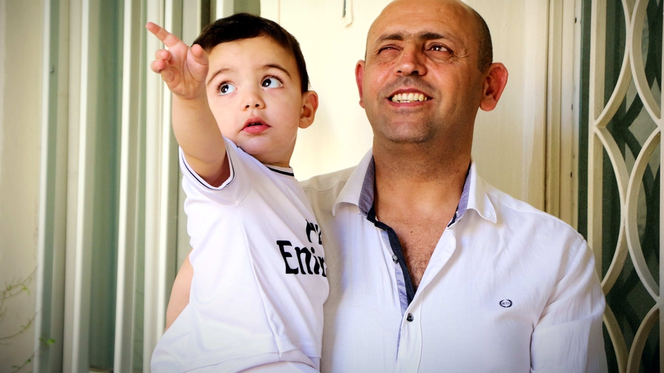 Elie smiles as he carries Mirna's toddler Chris, who points animatedly. Both in white shirts. "Welcome" hangs from the wall.