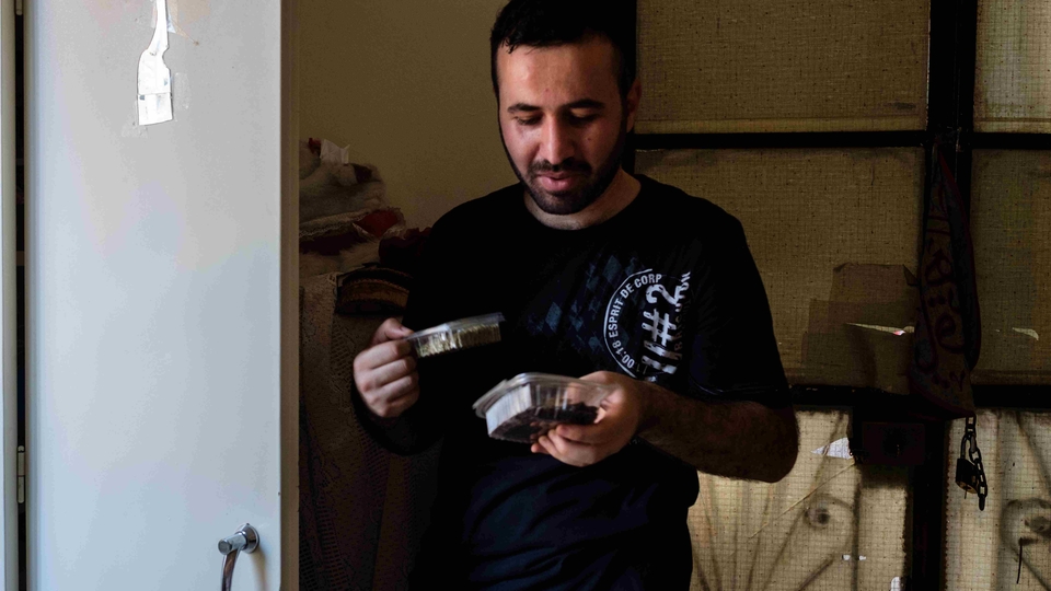 Mohammad Ali, dressed in a black Esprit De Corp T-shirt, looks down at two small plastic containers he is holding as he proudly describes their contents.