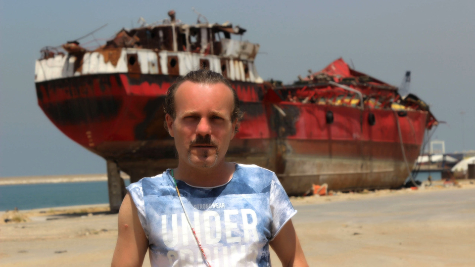 Portrait of Viken, in a blue and white T-shirt, standing in front of a red, destroyed ship docked at the port.