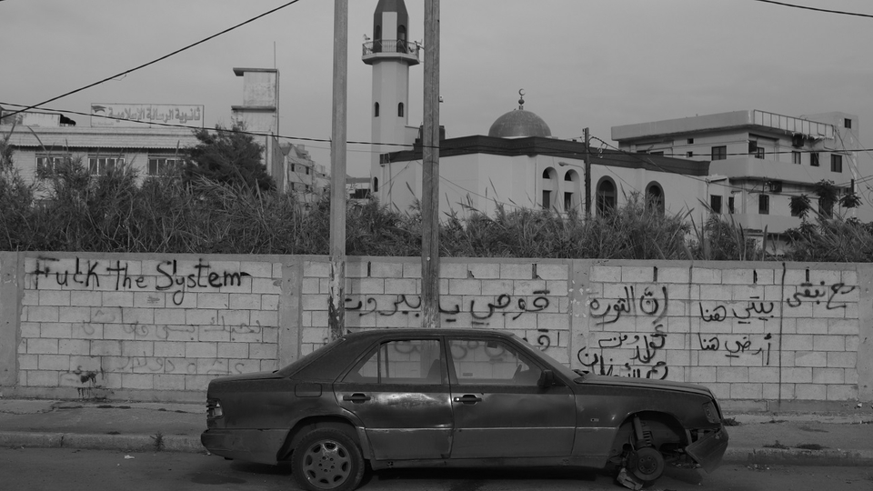 one wrote on the wall, “We will stay, my house is here, here, my land is here, rise Beirut, rise from under the rubble.” Parked by the wall is a car that has been there for a while, its front wheel likely stolen. To the left, one reads, “Fuck the system, I love you, but my salary is ten dollars,” signed by Abu Abeer.